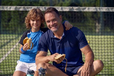 Memnuniyetle kişisel antrenör ve muzlu çocuk mahkemede padel antrenmanı sırasında ağların yanında otururken kameraya gülümseyerek bakıyor.