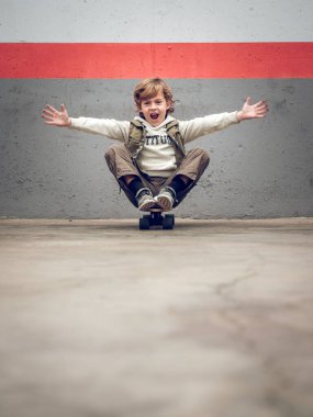 Moda kıyafetli, kollarını kaldırıp kaykayın üzerine oturmuş ağzı açık kameraya bakan neşeli küçük bir çocuk.