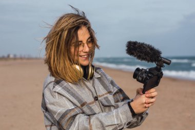 Sıcak kareli gömlekli neşeli genç bayan blogcu kumlu sahilde gülümserken bulutlu gökyüzüne karşı kamerada vlog kaydediyor.