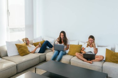 Modern dizüstü bilgisayarlar ve akıllı telefonlar üzerinde sörf yapan odaklanmış bayan arkadaşlar açık oturma odasında rahat bir kanepede otururken evde masa var.