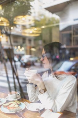 Moda kıyafetleri içinde güzel bir bayan elinde bir fincan sıcak içecekle ve rahat bir kafede pencerenin arkasında otururken başka tarafa bakıyor.