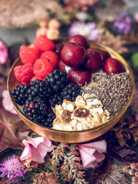 Kirazlı, ahududu ve böğürtlenli, mısır gevreğinin yanına yerleştirilmiş, çiçeklerle süslenmiş metal bir yoğurt kasesinden.