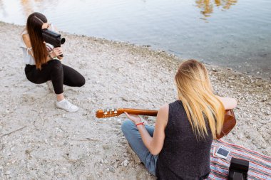 Gündelik kıyafetli genç bayan, plajda birlikte vakit geçirirken, isimsiz bir bayan arkadaşının akustik gitar çalışını çekmek için kamera kullanıyor.