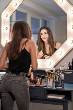 Makyajlı, uzun siyah saçlara dokunurken aynada ampullerle yansımaya bakan zayıf bir kadın.