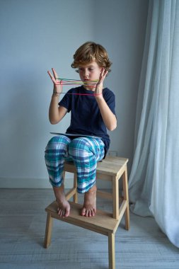 İlginç kediler oynayan, odaklanmış bir çocuk. Işık odasındaki tabletle tahta taburede otururken beşik oyunu oynuyorlar.