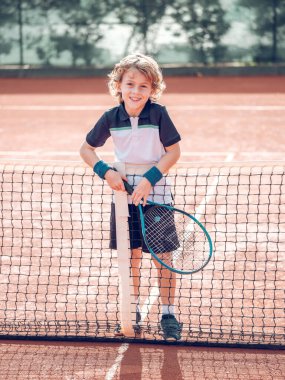 Aktif giyimli, kıvırcık sarı saçlı, elinde raketle tenis kortunda kameraya bakarken gülümseyen ve kameraya bakan bir çocuk.