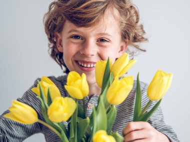 Kameraya gülümseyen iyimser çocuk, stüdyoda beyaz arka planda yeşil yapraklı renkli lalelerin yanında gülümsüyor.