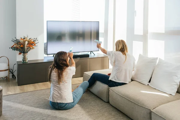 Back view of unrecognizable female friends with light pistols playing videogame on modern TV in light living room at home