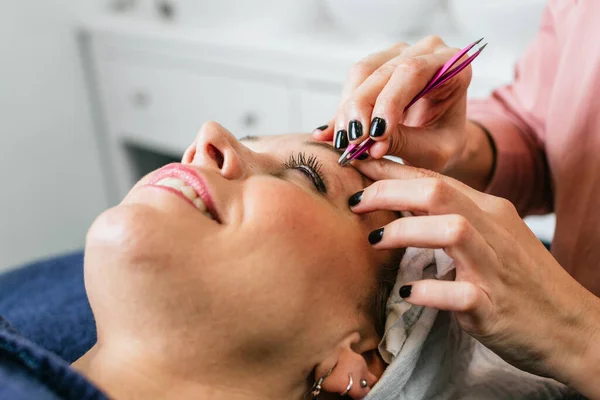 Cropped anonymous cosmetologist plucking eyebrows on face of female client lying on table in beauty salon