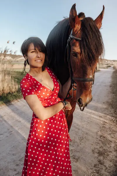 Kırsal kesimde güneşli bir günde çiftliğin avlusunda dikilirken uzun kırmızı elbiseli genç bayan kameraya bakıyor.