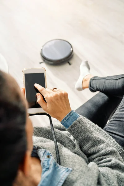 Tanımlanamayan bir kadının kanepeye oturması ve akıllı telefonlara göz atması, modern robotik elektrik süpürgesinin evdeki oturma odasında yerde çalışmasıdır.