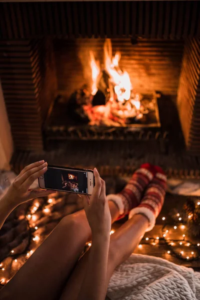 スマートフォンを使用して暖かい靴下の認識できない女性は居心地の良い部屋の暖炉の近くに座っている間燃える火の写真を撮ります — ストック写真