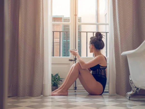 ホテルのバスルームでコーヒーを飲みながら窓の近くに座っている黒いボディースーツのスリムな官能的な裸足の女性の側面 — ストック写真
