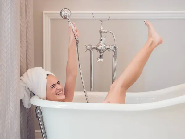 快乐的女性 头戴毛巾 躺在烧杯浴缸里 愉快地举起手臂 一边用水龙头和腿 一边玩乐 — 图库照片