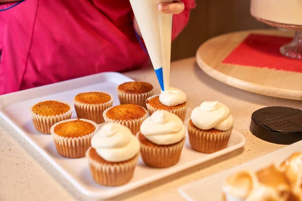 Unrecognizable crop person decorating cupcakes using piping bag standing at kitchen counter with other desserts on it