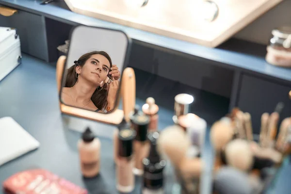 Crop anonymous master applying cosmetic product on face of female client reflecting in mirror at table with makeup supplies in beauty salon