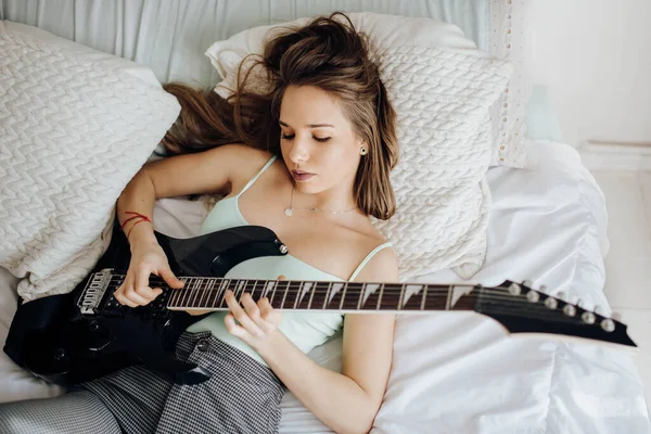 上から若いブルネットの女性がベッドの上でギターを弾く — ストック写真
