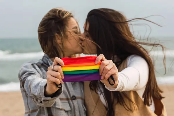 多云的日子里 穿着休闲装的年轻夫妇在沙滩上接吻时 双目紧闭 面膜上挂着Lgbt彩虹旗 — 图库照片