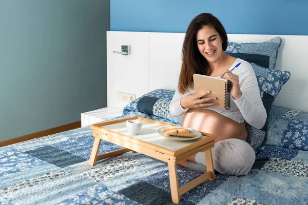 Ciężarna Kobieta Siedzi Łóżku Pisze Notatniku Podczas Śniadania Rano — Zdjęcie stockowe