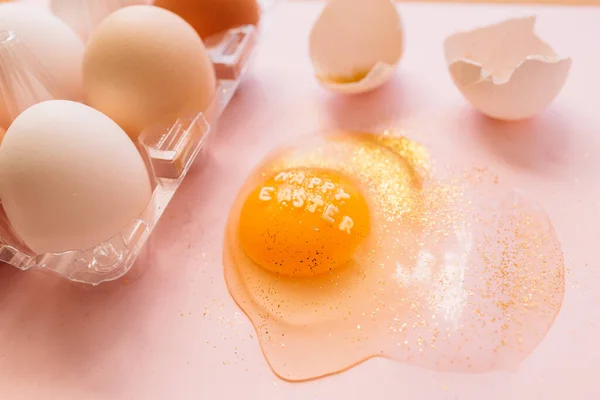 テキスト付き壊れた輝く卵のトップビューピンクの背景にプラスチック製の箱にハッピーイースターと生の卵 — ストック写真