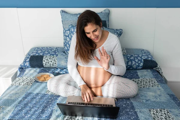 从上图中可以看出 孕妇一边坐在舒适的床上一边一边一边用电视在上网本打电话 一边挥手 — 图库照片