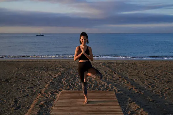 Full body of Hispanic female in sportswear doing Tree pose with namaste hands during yoga session on sandy seashore near sea