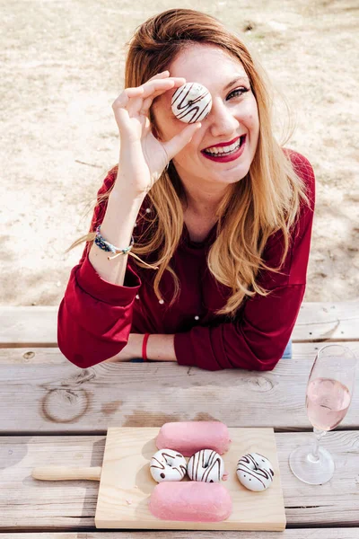 年轻迷人的微笑女人坐在野餐桌旁 透过甜甜圈的孔望过去 — 图库照片