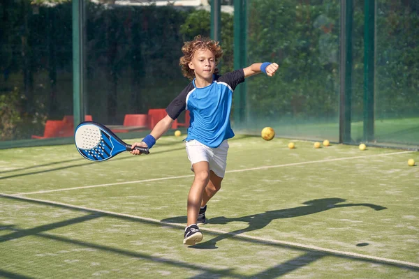Aktif giyimli bir çocuk yaz günü spor sahasında Padel oynarken raketle topa vuruyor.