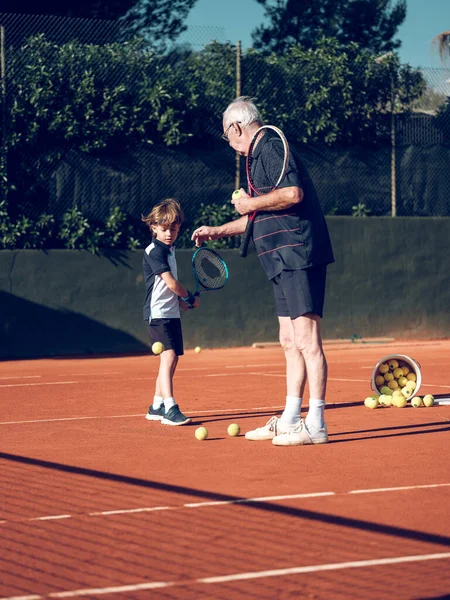 Full body of focused senior instructor explaining preteen player technique of backhand stroke with tennis racket while training in hard court