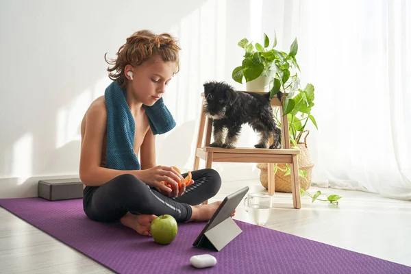 Shirtless boy sitting with crossed legs on yoga mat and listening to music from tablet while eating fruits near dog