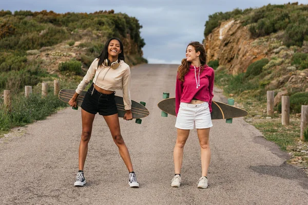 Full body of happy multiethnic girlfriends standing on road with longboards