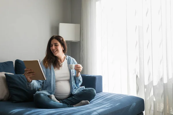 Positiv Schwangere Frau Sitzt Auf Sofa Mit Notizbuch Und Trinkt — Stockfoto