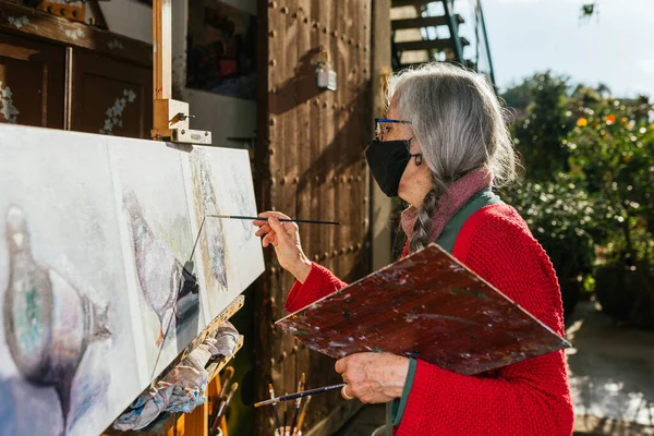 Side view of aged female artists in protective mask painting on canvas in backyard on sunny day in summer