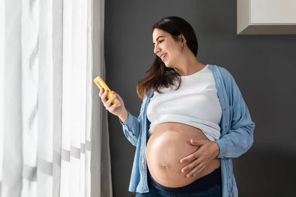 Smiling Pregnant Female Touching Belly While Standing Room Home Browsing — Stok fotoğraf