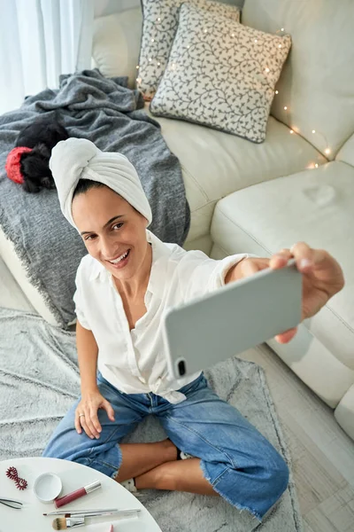 以上是一位穿着浴巾头巾 快乐的年轻女性选美博主在智能手机上为博客内容自拍的故事 她坐在地板上 双腿交叉 桌子上摆着化妆品 — 图库照片