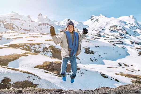 Happy kid wearing warm clothes and hat with mittens jumping high with raised arms near snowy hills under blue sky and looking away in sunny winter day