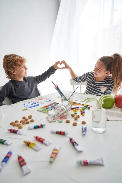 Optimistic children making heart gesture and looking at each other while sitting at table with various paints and colorful drawings