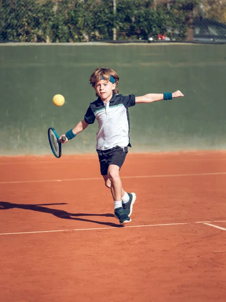Spor giyim ve saç bandı giymiş, güneşli bir günde kortta raketle tenis oynayan küçük bir çocuğun tüm vücudu.