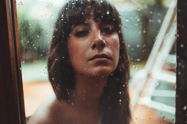 Beautiful female behind wet surface transparent glass and looking at camera