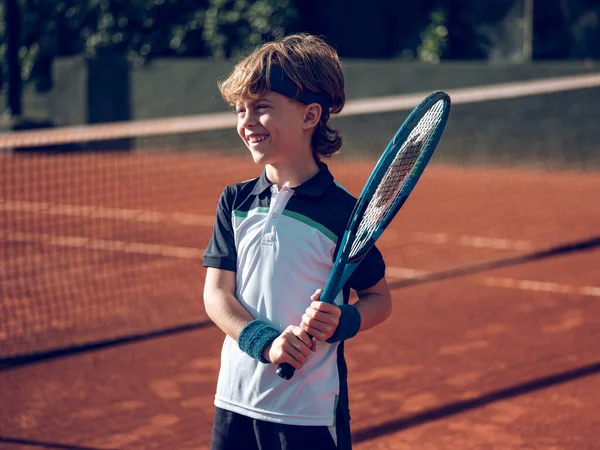 Güneşli bir günde antrenman yaparken profesyonel tenis raketiyle sahada gülümseyen çocuk.