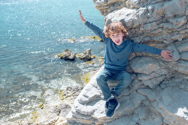 上からポジティブな男の子の完全な体から 海の上の石の崖の上に座っている間 カメラを見ている広がりの腕 — ストック写真