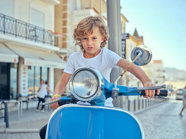 Kıvırcık saçlı çocuk, Portekiz 'in Nazare şehrinde açık yaz gününün tadını çıkarırken mavi scooter sürerken komik bir ifade takınıyor.