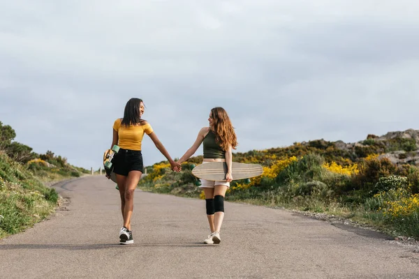 Full body of happy multiethnic girlfriends holding hands looking at each other while walking on road with longboards