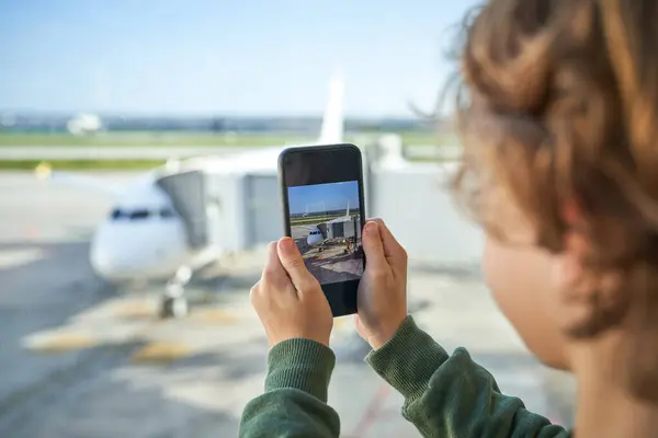 Havalimanı terminalinin panoramik penceresinin yanında duran modern uçağın fotoğrafını çekerken günlük kıyafetleri içinde sarı saçlı tanınmayan küçük çocuk akıllı telefon kullanıyor.