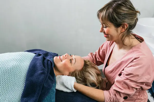 Side view of cheerful beautician wiping face of female client with towel during skin care routine in spa salon