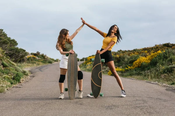 Full body of happy multiethnic girlfriends giving high five while standing on road with longboards
