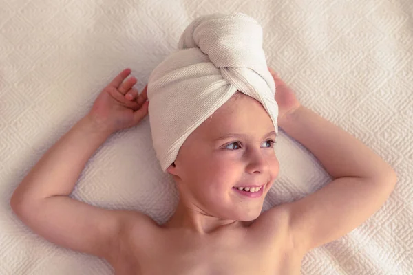 Top view of happy little boy with towel on head and naked torso resting on white blanket with raised arms and looking away