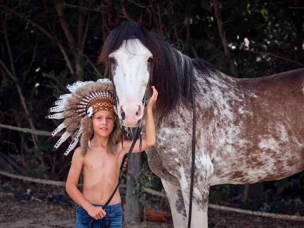 stock image Positive preteen boy in traditional indigenous North American headgear caressing obedient roan horse with reins and bridle and looking at camera while standing near fence and trees in countryside