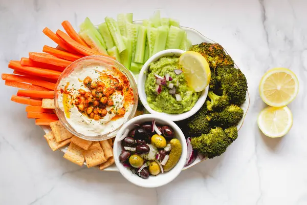 İştah açıcı sağlıklı humus yeşil sosu havuç, salatalık ve yeşil brokolili salatalık dilimlenmiş limon dilimlenmiş beyaz kaseler ve masada.