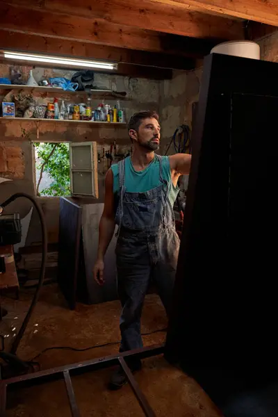 Kot giyen ciddi sakallı bir erkek işçi hafif atölyede çeşitli ekipmanlarla ayakta duruyor ve iş sırasında pencereyi açıyor.